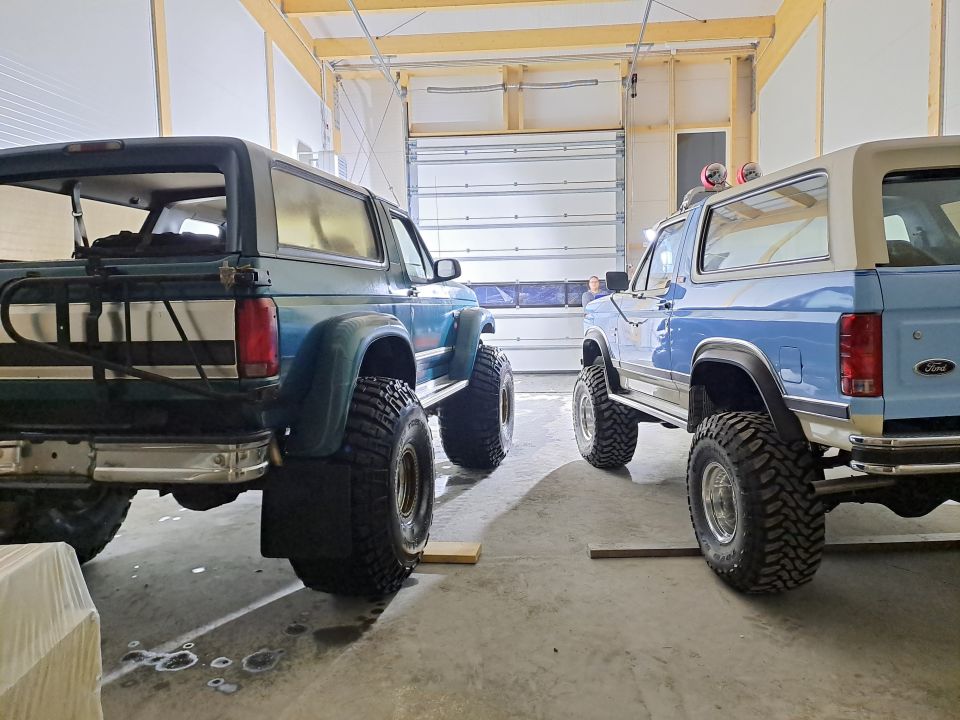 Two lifted Ford Broncos, one dark green and one light blue, parked in a garage.