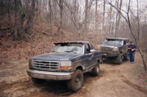 1992 Ford Bronco - Two Ford Broncos navigate a wooded trail, kicking up dust on the muddy path.