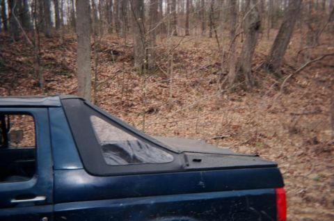 1992 Ford Bronco - A dark blue SUV with a soft top over its rear cargo area is parked in a wooded area.