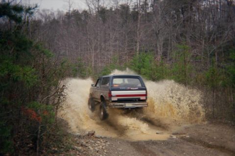 1992 Ford Bronco - A 1992 Ford Bronco splashes through a muddy puddle on a dirt road surrounded by trees.