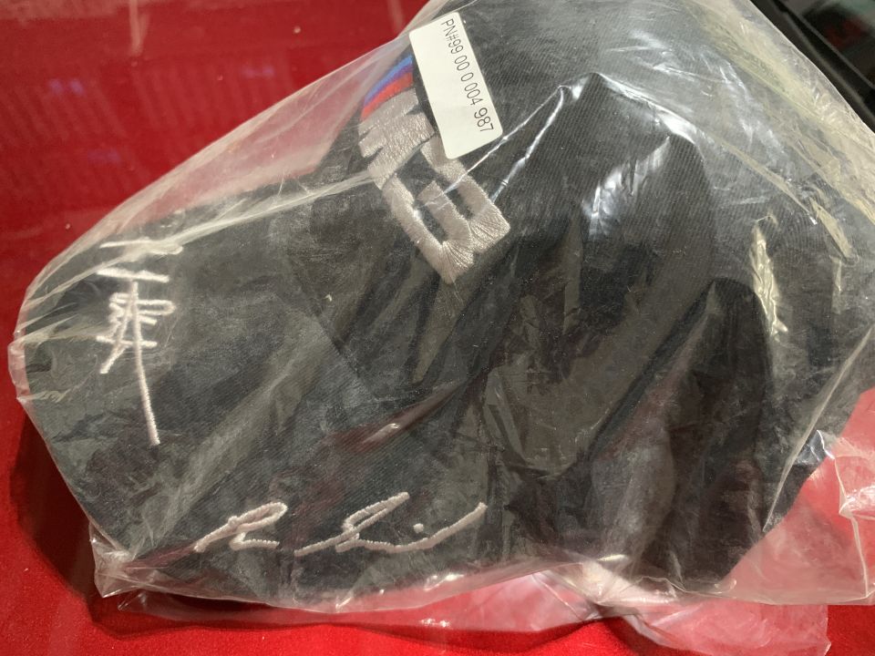 Black baseball cap in plastic bag with embroidered text and part number label.