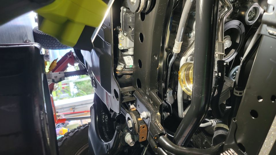 Underside view of vehicle engine bay with hoses, pulleys, and a yellow jack.
