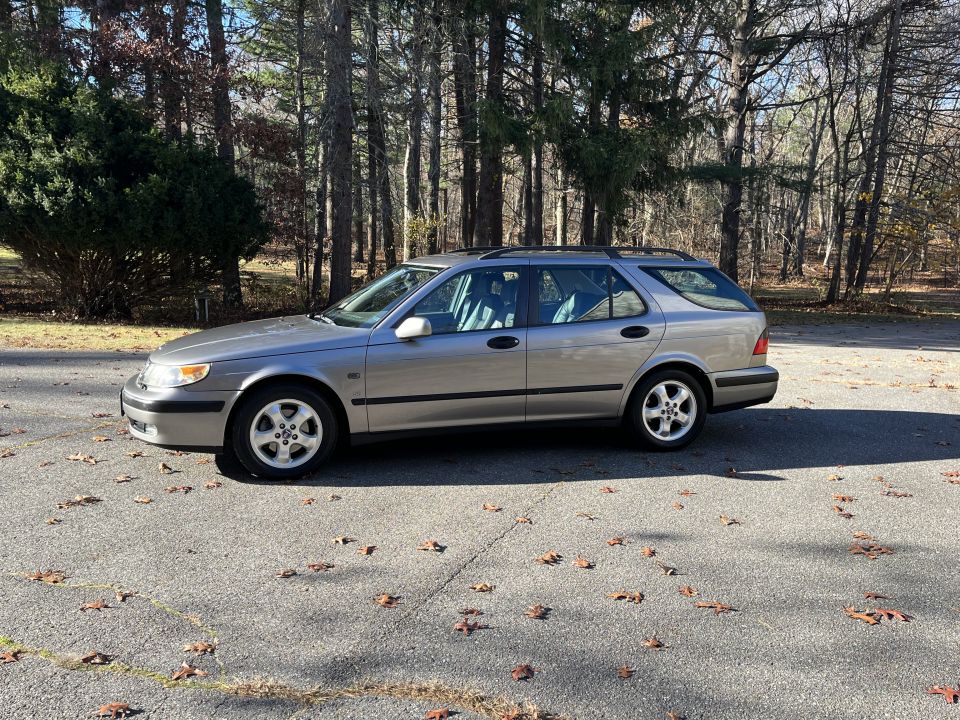 Silver wagon parked on asphalt with fallen leaves. The vehicle has a roof rack and is viewed from the side