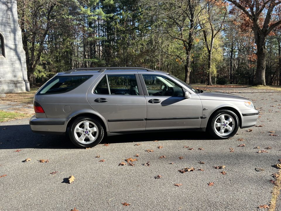Side view of a silver wagon parked on asphalt with fallen leaves. Trees and a stone structure are in the background