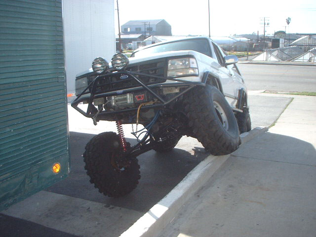 White lifted Ford Bronco flexing front suspension off a curb, tube bumper with winch and round off-road lights