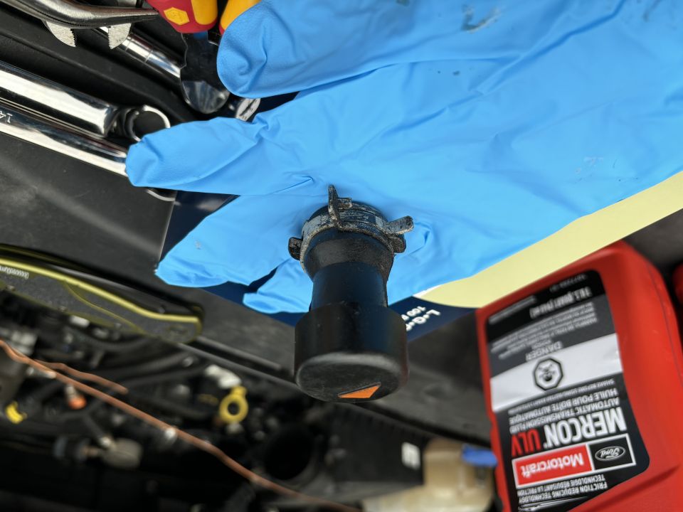 Blue gloved hand holds radiator cap over engine bay with red MERCON ULV fluid bottle nearby