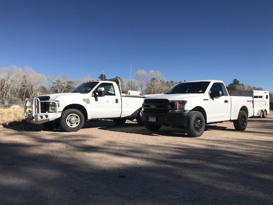 Two white Ford pickup trucks parked on a dirt road, one larger with a custom bumper, the other newer towing a trailer.