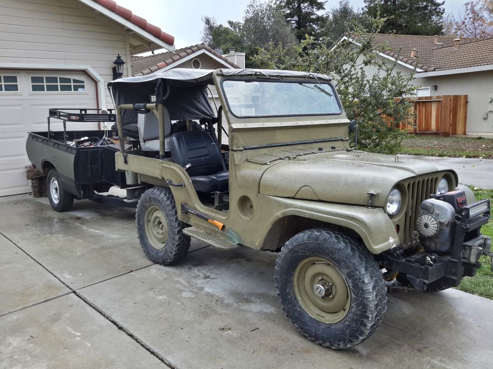 1952 Jeep CJ5 with a soft top, towing a small utility trailer. The Jeep has an aftermarket bumper with a winch and lights