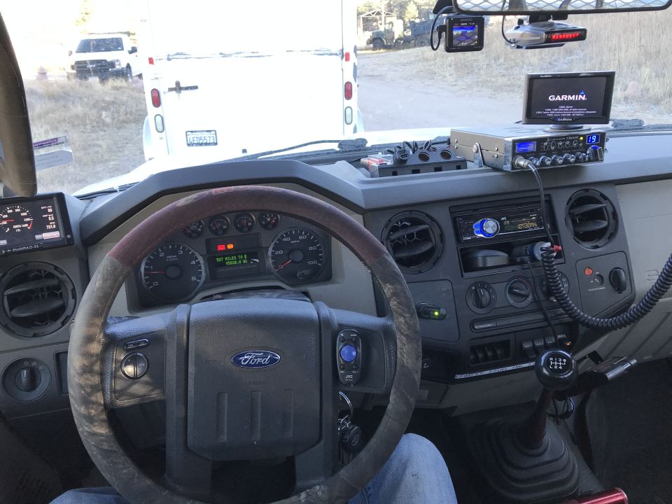 Interior of a truck cab, showing steering wheel, dashboard, CB radio, and Garmin GPS. Another truck is visible outside