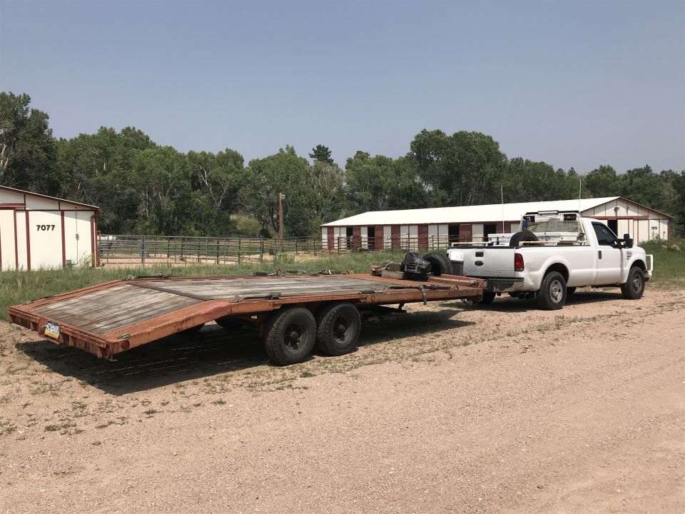 White Ford F-250 pickup truck towing a flatbed trailer near a barn.
