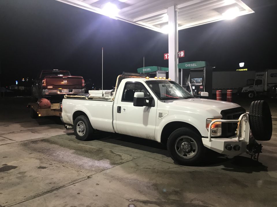 White Ford F-250 towing a red pickup truck at a gas station at night.