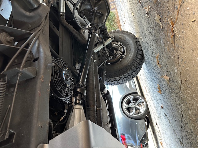 Undercarriage of lifted Bronco showing driveshaft, rear axle, electric fan, and off-road tire; gray Audi in background