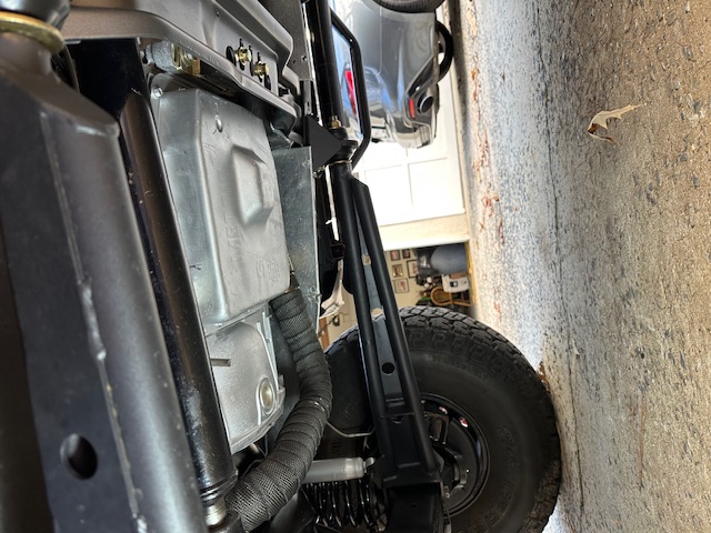 Undercarriage of a Ford Bronco showing aluminum pan, wrapped exhaust, black sliders, and all-terrain tire in a garage