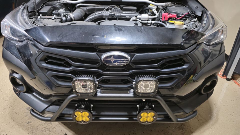 Black Subaru Impreza front view with bull bar and auxiliary lights, hood open.