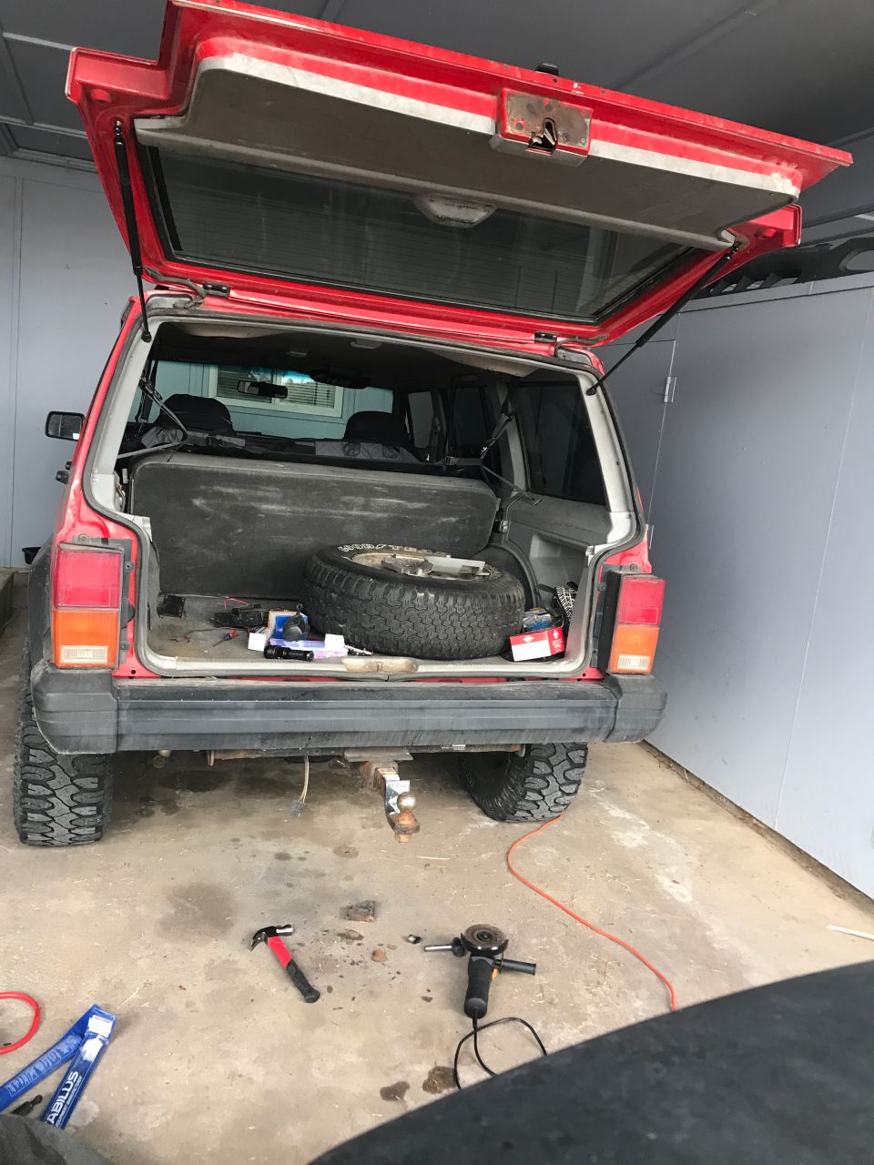 Red Jeep Cherokee rear view with tailgate open, spare tire inside, tools on ground.