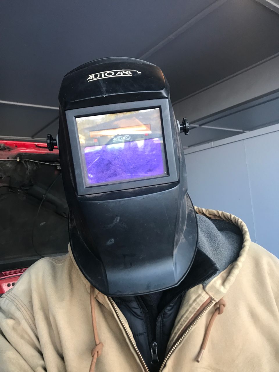 Person wearing a welding helmet in front of an open red vehicle hood.