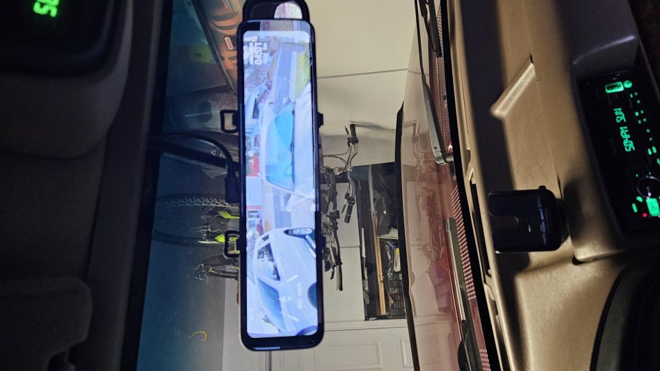 Smartphone mounted on vehicle dashboard showing backup camera feed of a white car and bicycles.