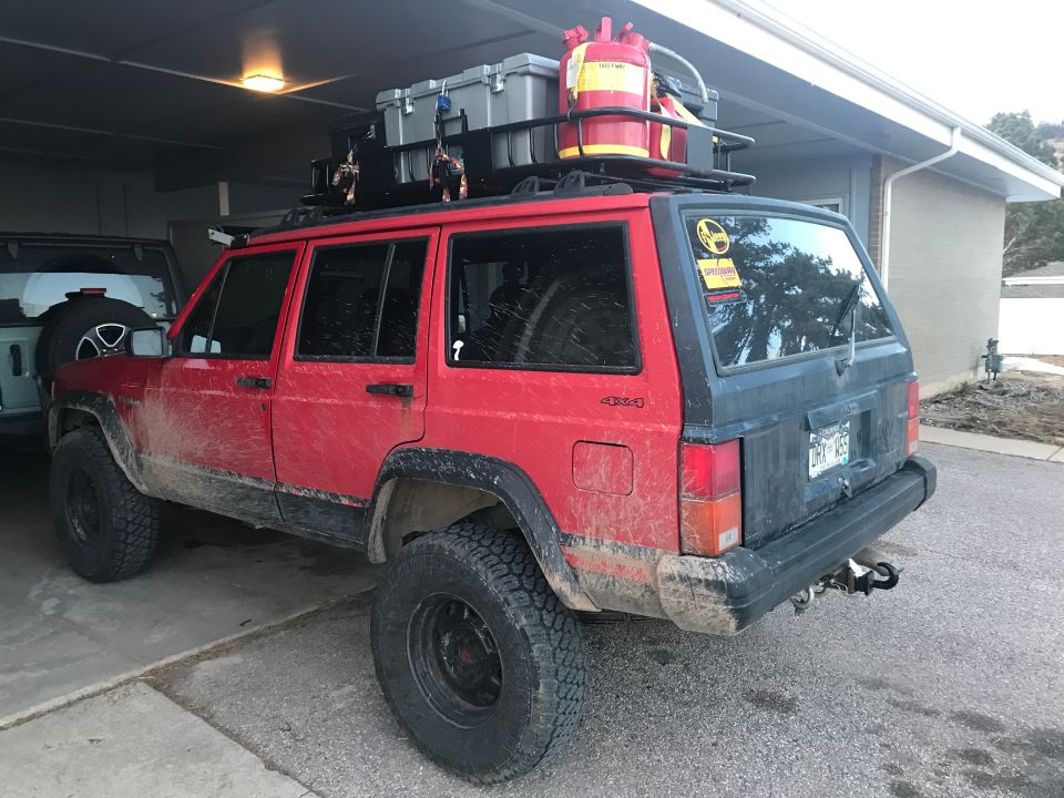 Red and black 1996 Jeep Cherokee with roof rack, gas cans, and storage boxes, covered in mud.