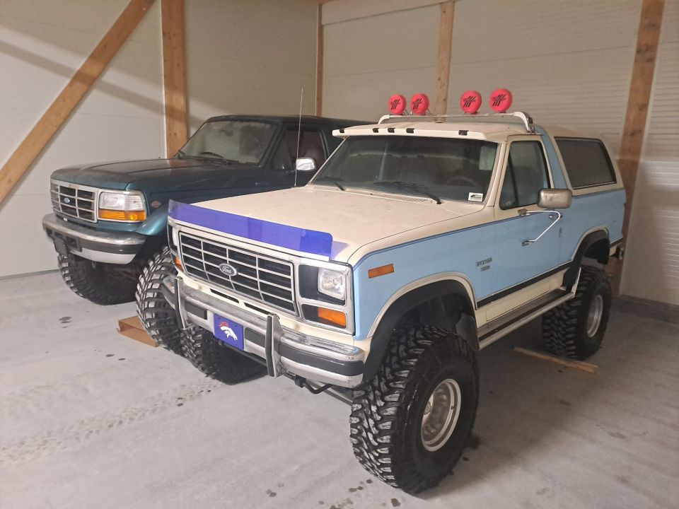 Two-tone blue and white Ford Bronco with off-road tires and roof lights, parked next to a dark green Ford F-Series truck