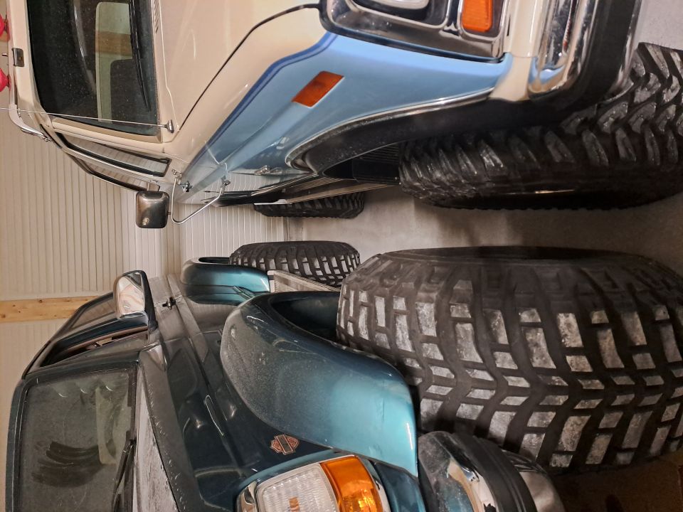 Teal vehicle and white vehicle with blue stripe parked closely; large off-road tires visible