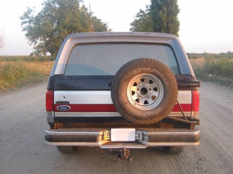 1988 Ford Bronco - Rear view of a dark blue and silver SUV with a spare tire on the back, parked on a dirt road.