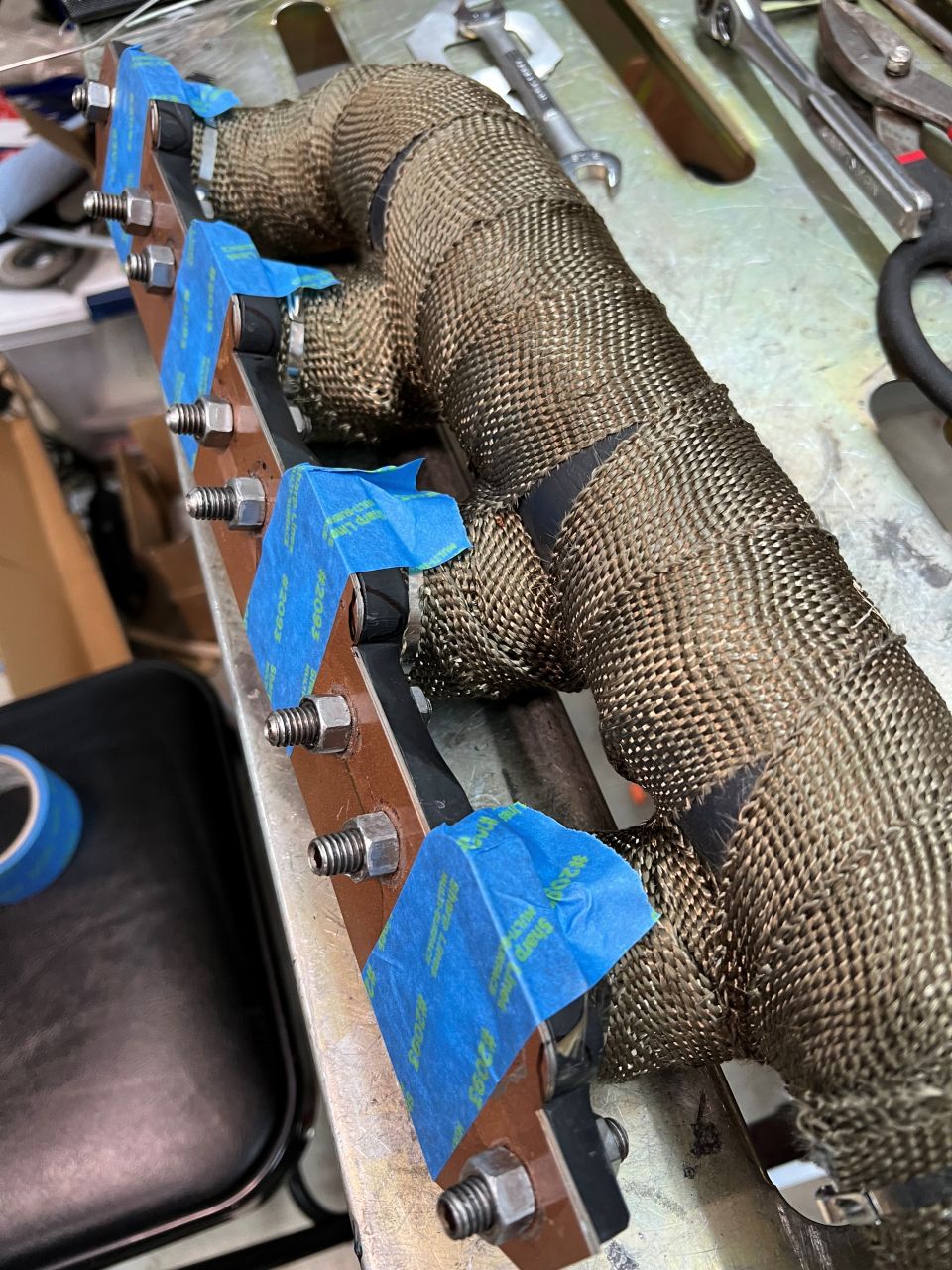 Custom exhaust manifold wrapped in heat wrap, secured with blue tape, on a workbench.