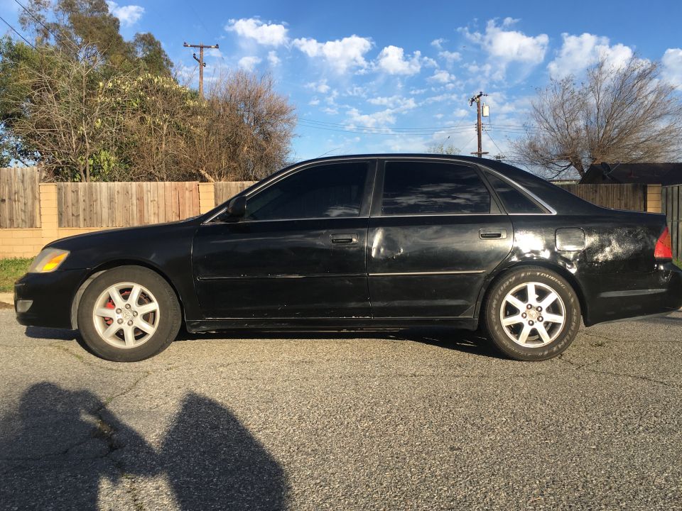 Black sedan in side profile parked on street by a wooden fence, with alloy wheels and dented doors