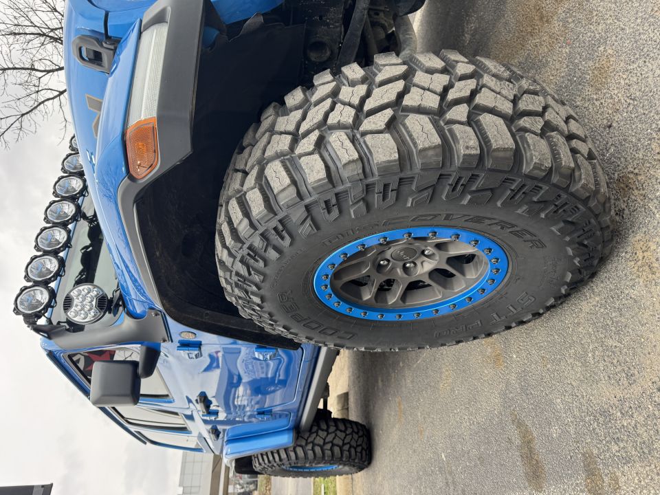 Blue Jeep Wrangler with large off-road tires and auxiliary lights, close-up on front passenger side.