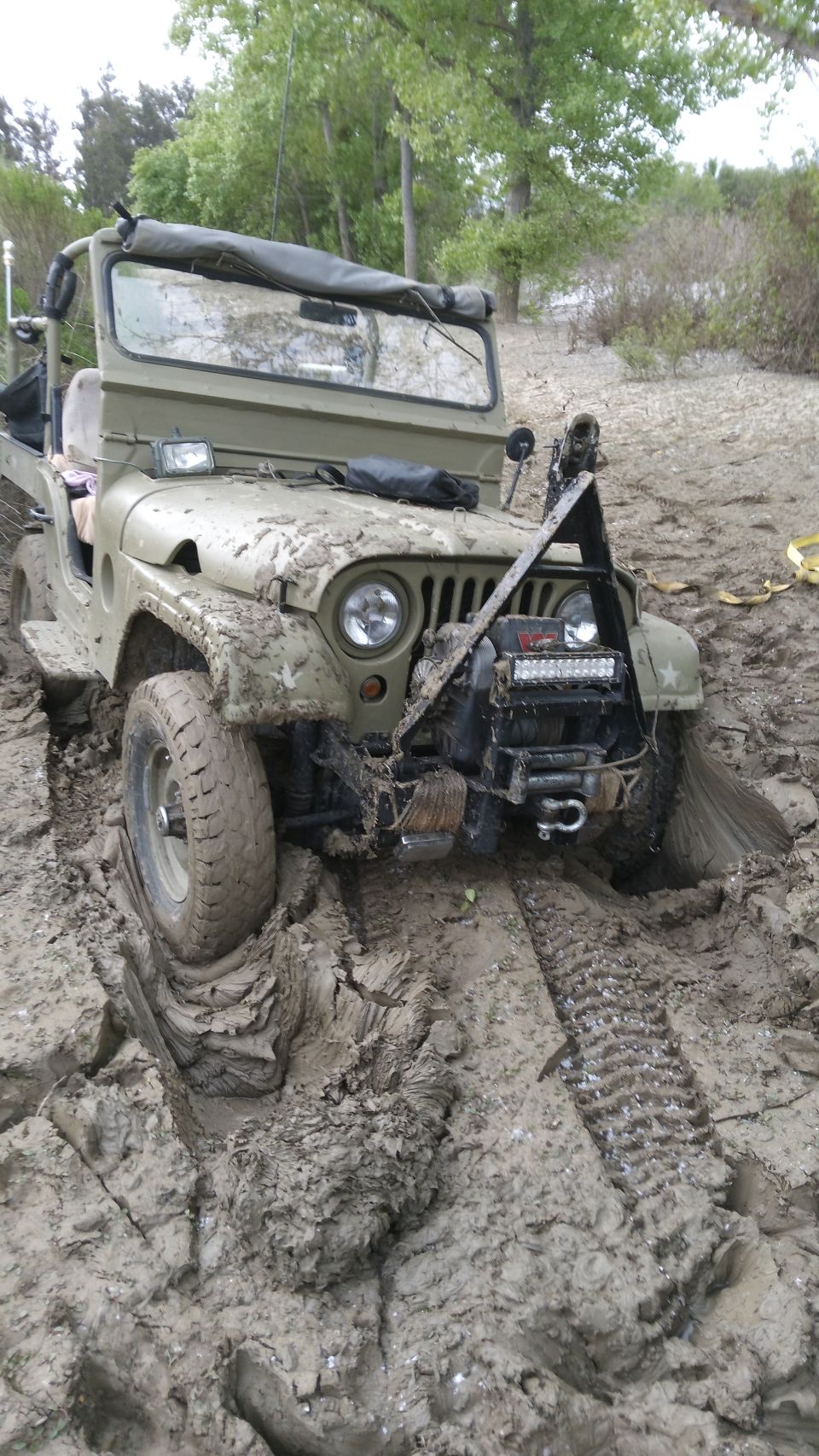 Mud-covered olive green Jeep CJ5 stuck in mud, front tire embedded, winch and lights visible.