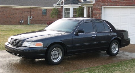 1999 Ford Crown Victoria - A dark-colored sedan is parked on a wet driveway in front of a residential building.