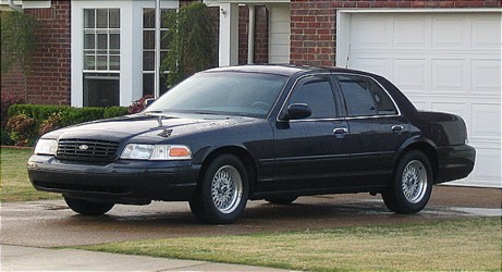 1999 Ford Crown Victoria - A black sedan is parked in front of a brick house and a garage door.