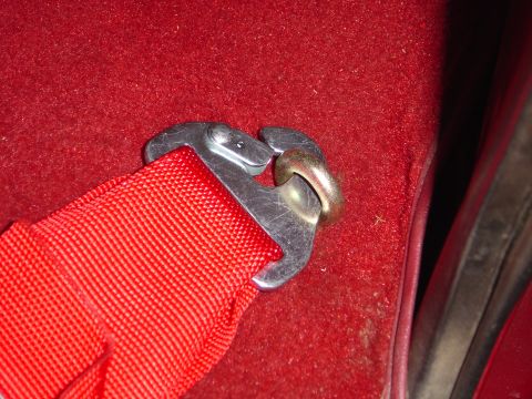 1978 Ford Bronco - Close-up of a red racing harness belt with a steel snap hook clipped to a floor-mounted eye bolt, set into red carpet beside a body seam an…