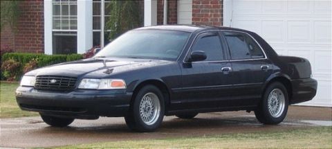 1999 Ford Crown Victoria - A dark-colored sedan is parked in front of a residential garage and brick house.