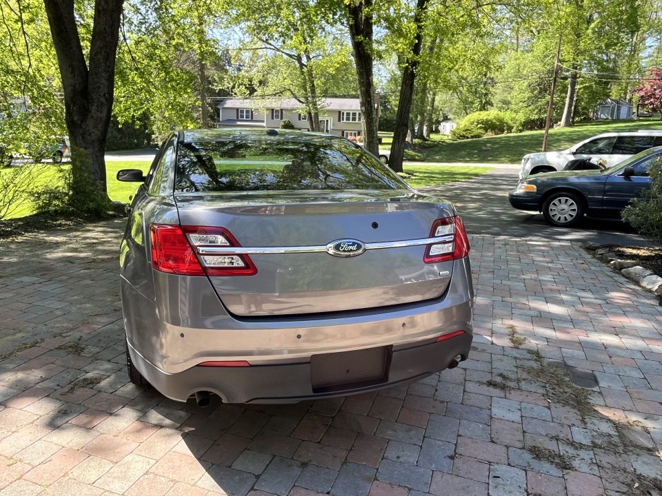 Rear view of a gray Ford Taurus parked on a brick driveway. Another car is visible to the right