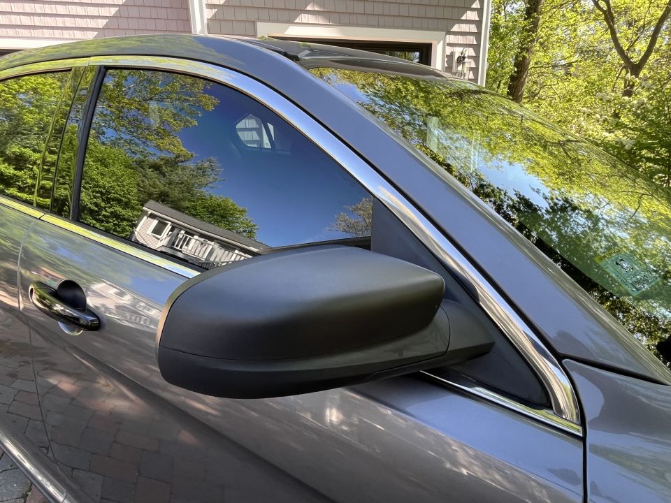 Side view of a gray SUV parked in front of a house. Reflections in the window show trees and the house