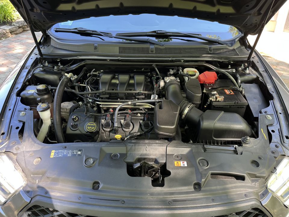 Engine bay with V6 engine, air intake, battery, and various hoses visible