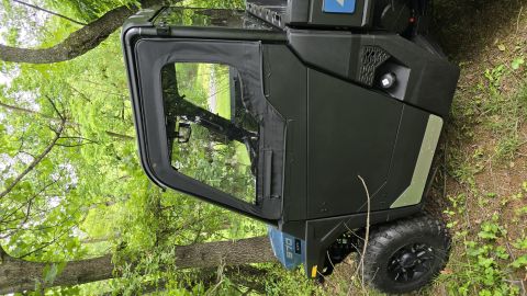 2025 Polaris RANGER - A dark Polaris UTV is parked outdoors, partially obscured by trees and foliage. A '570' badge is visible on the rear panel.