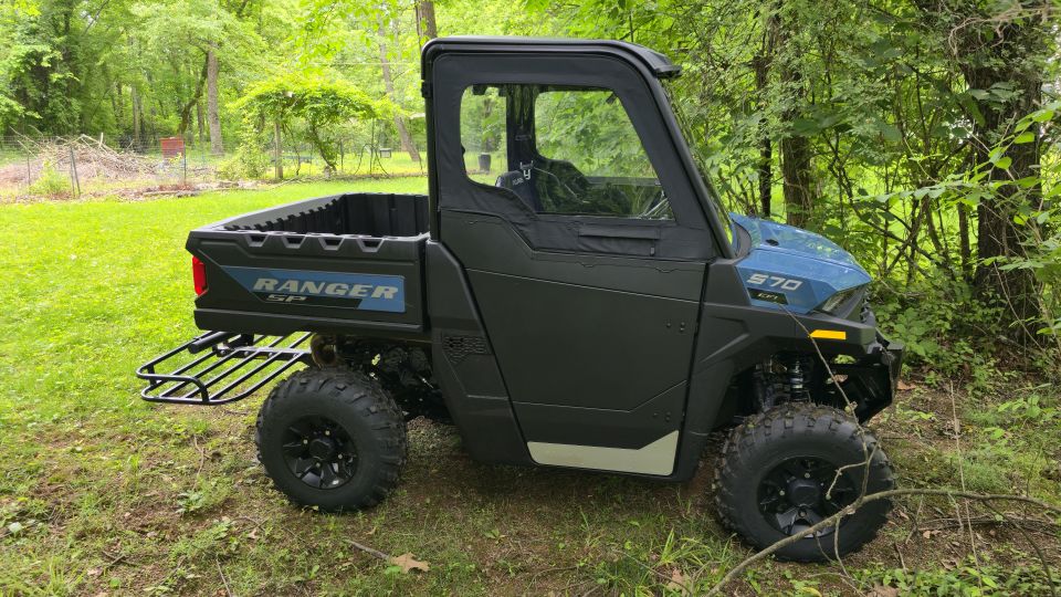 Side view of a blue Polaris Ranger UTV with a black cab and bed, parked in a grassy, wooded area. It has a rack on the back