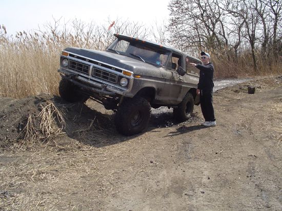 Dark gray 1978 Ford Bronco on a dirt path, angled up a small incline, with a person standing beside it