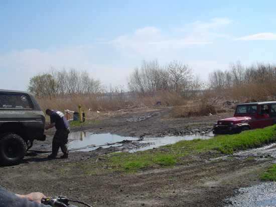 Dark SUV and red Jeep in muddy terrain with a person working on the SUV