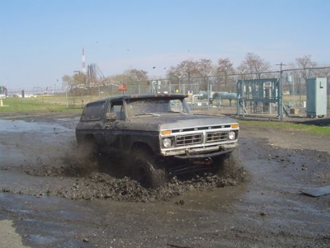 1979 Ford Bronco - This 1978 Ford Bronco is navigating a challenging muddy terrain with industrial fencing and structures visible in the distance.