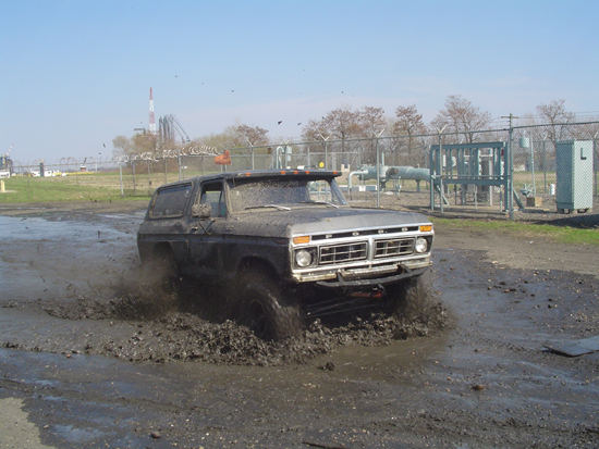 Ford Bronco driving through a mud pit, kicking up water and dirt. Industrial structures in background