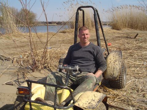 1979 Ford Bronco - A man is seated in a yellow dune buggy, parked on a sandy bank near dry reeds and water.
