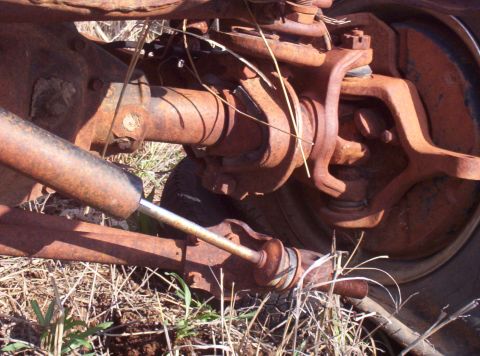 1988 Ford Bronco - Close-up view of heavily rusted front axle and suspension parts, including a shock absorber, set against dry grass.