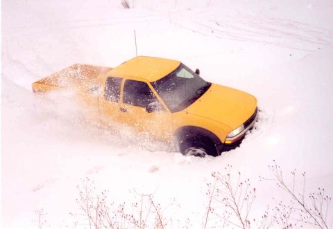 1995 Ford Bronco - A yellow pickup truck is stuck in the snow, with snow spraying around its tires.