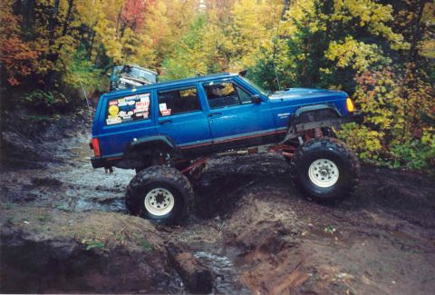 1995 Ford Bronco - A lifted blue Jeep Cherokee XJ tackles a challenging muddy trail during autumn, with another vehicle visible in the background.
