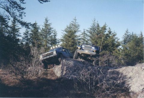 1995 Ford Bronco - Two off-road vehicles navigate a rocky terrain with trees in the background.