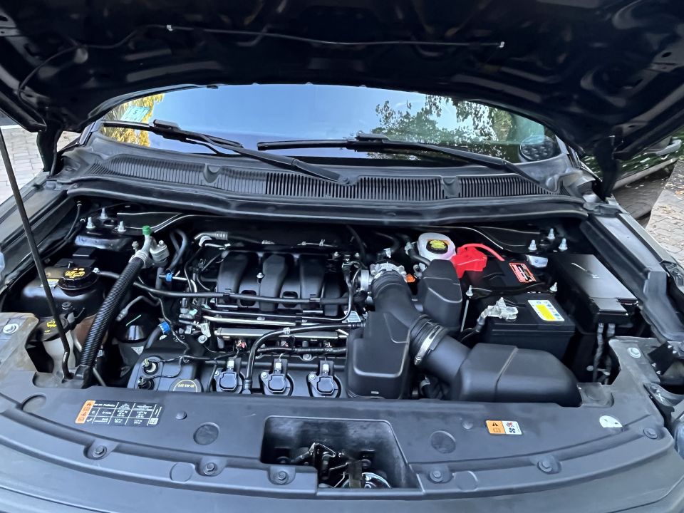 Engine bay of a black vehicle with the hood open, showing the engine, battery, and various hoses and wires