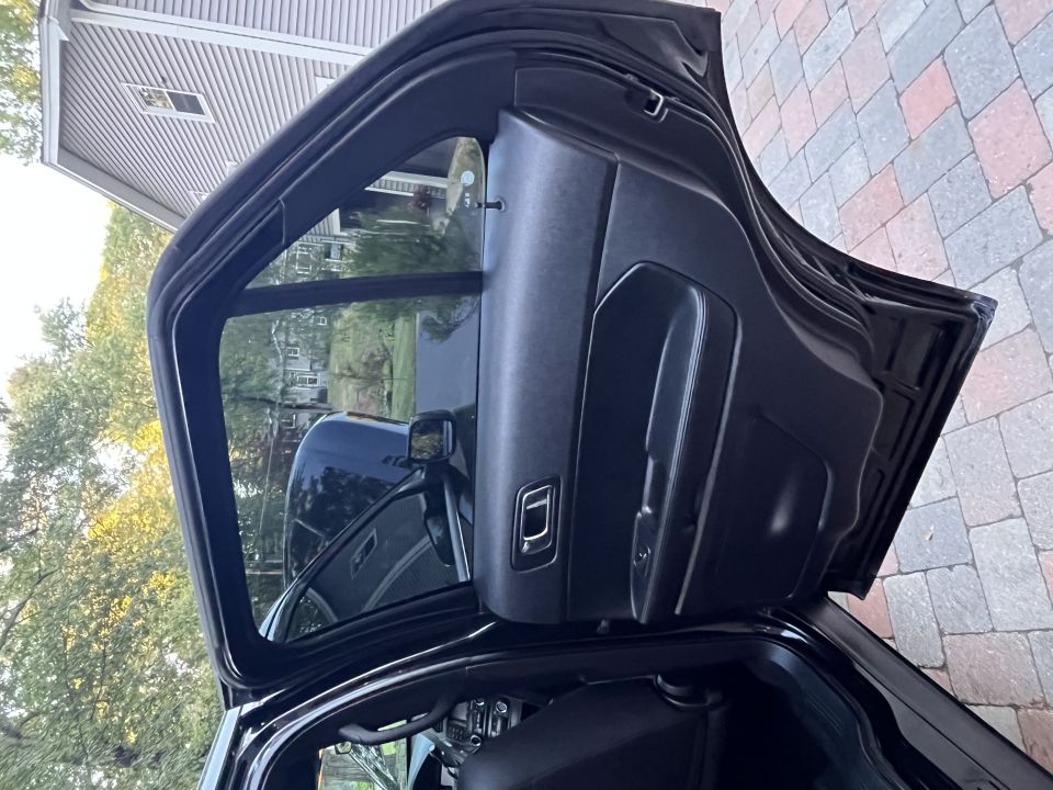 Black car interior, driver's side door open, showing door panel and dashboard. Paved driveway visible