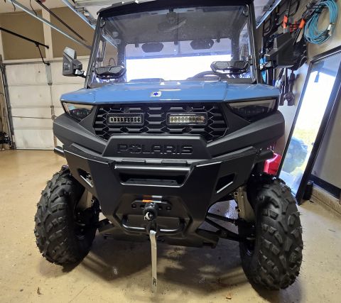 2025 Polaris RANGER - A blue Polaris UTV is parked indoors, showing its front grille, bumper, and headlights. The vehicle has a winch attached to the front bumpe…